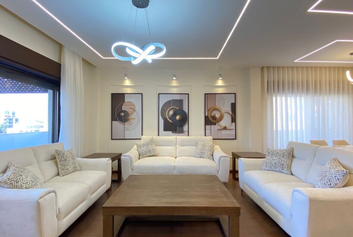Bright modern living room with white sofas arranged around a wooden coffee table and framed abstract art on the wall, under decorative ceiling lights.