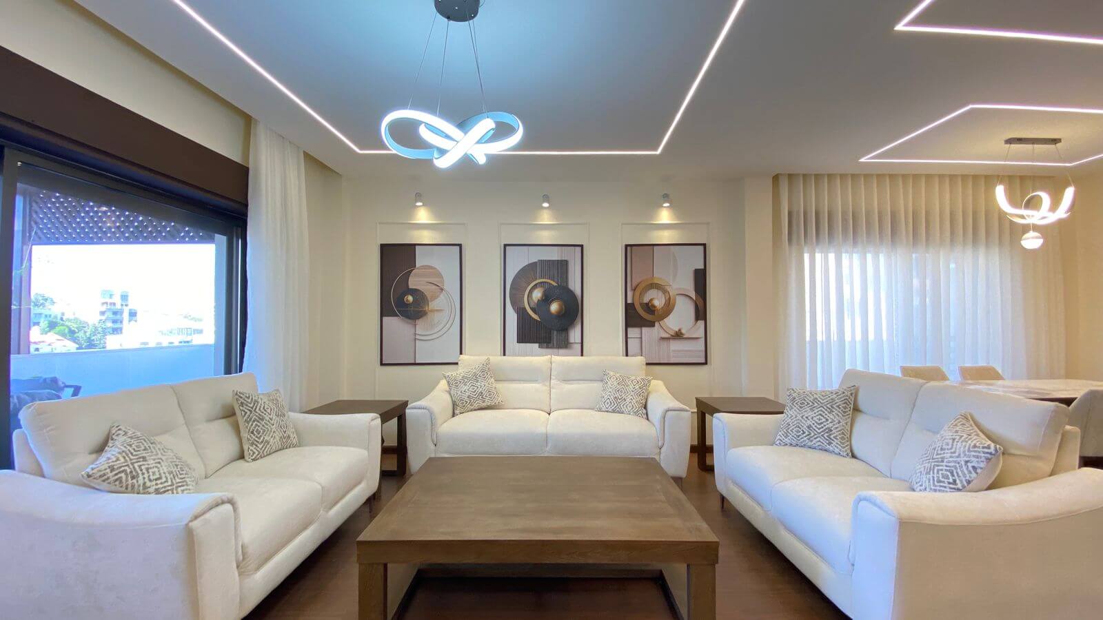 Bright modern living room with white sofas arranged around a wooden coffee table and framed abstract art on the wall, under decorative ceiling lights.