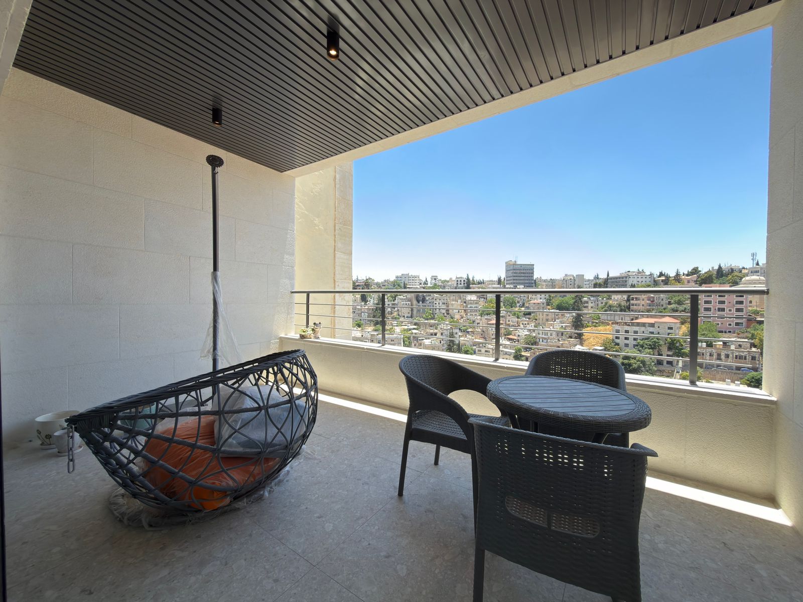 09e453d6-29d7-4028-8845-70e615966229 Balcony seating area with a round wicker table and four chairs, overlooking a cityscape under a blue sky.
