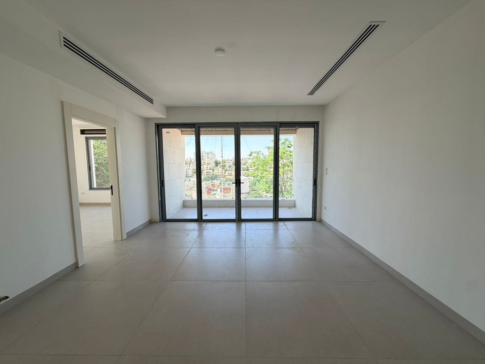 0b29986d-9ecd-4be6-9e62-fa6a5580eaa0 Bright empty living room with large glass sliding doors opening to a balcony overlooking a city, white walls and gray tiled floor.