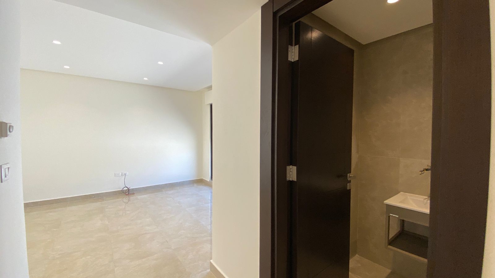0bdf503b-b7b0-4041-8337-962272abe241 Empty living room with beige tile floor, off-white walls, and recessed ceiling lights; dark doorway opens to a small bathroom with a sink.
