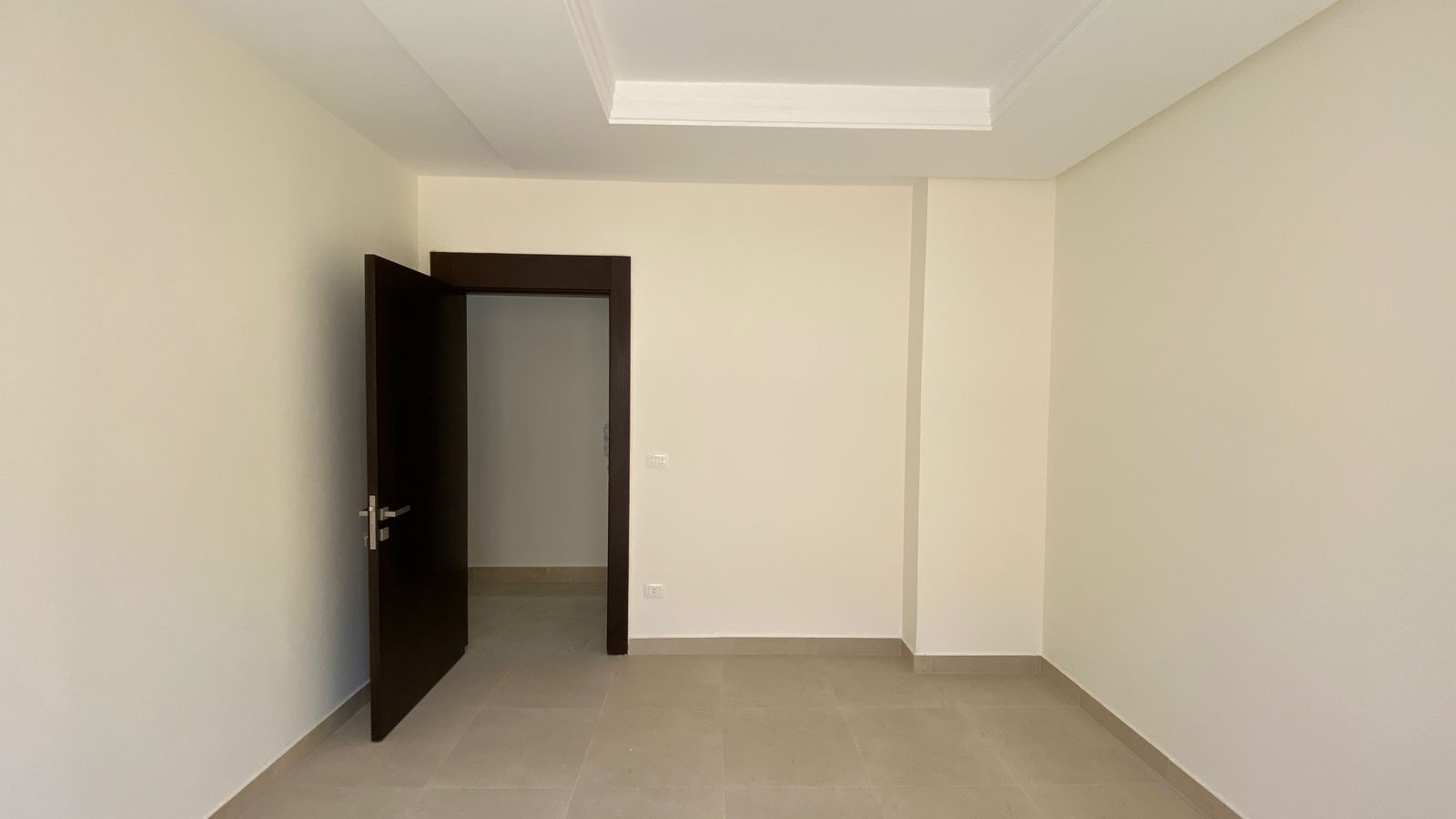 0cddedc4-94ff-496d-87e1-7bc8e99f73fe Empty room with beige tile floor, pale walls, and a dark wooden door ajar to another space. Ceiling light fixture visible.
