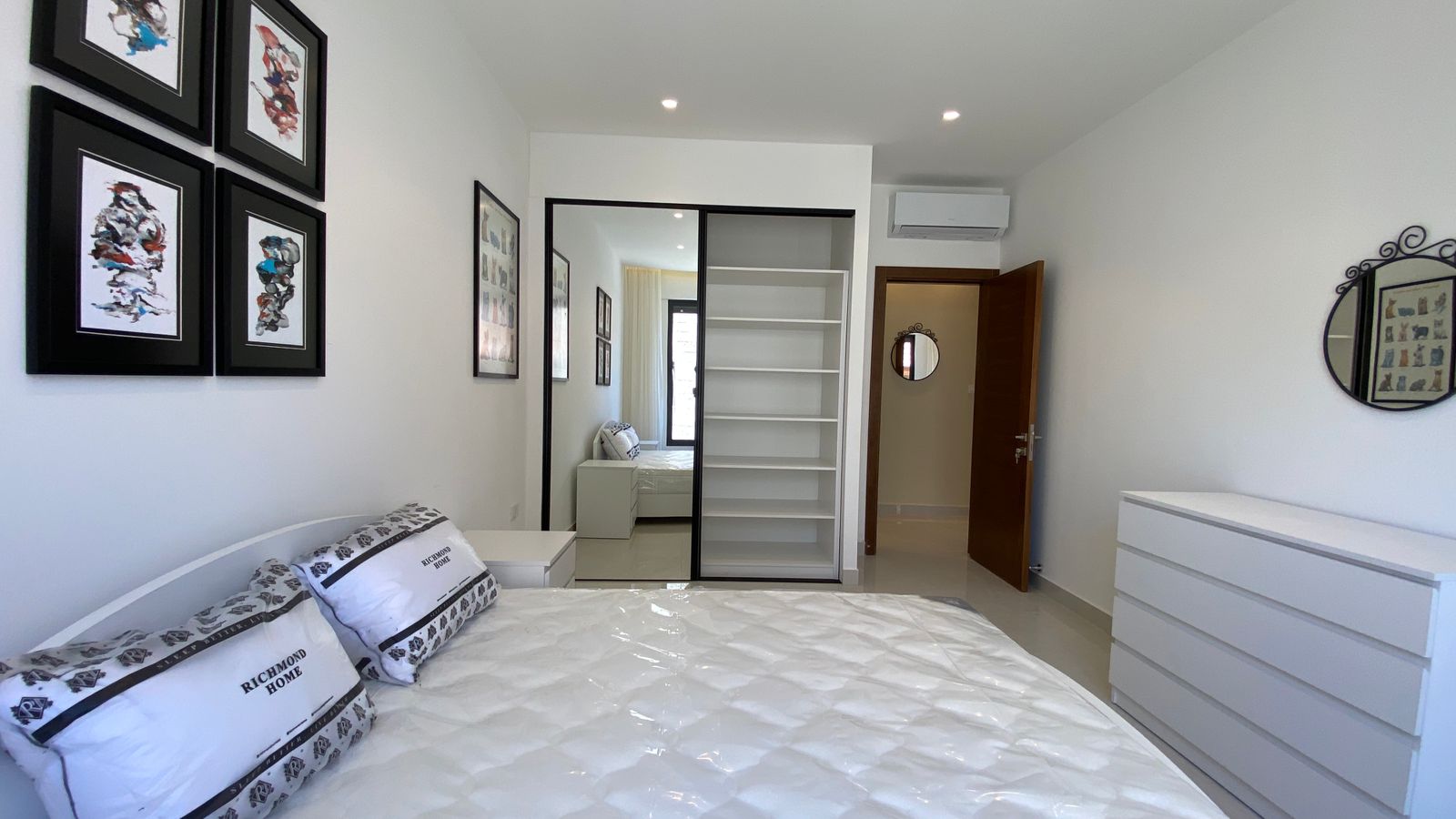 0dc6b4ca-9fc5-4fea-aec8-7e61524b501d Bright modern bedroom with white walls and a white bed; four black-framed artworks on the left wall; a mirrored sliding-door wardrobe with open shelves opposite; a white dresser on the right; a round decorative mirror above the dresser; ceiling recessed lights and a wall-mounted air conditioner near the door; a window with sheer curtains in the back