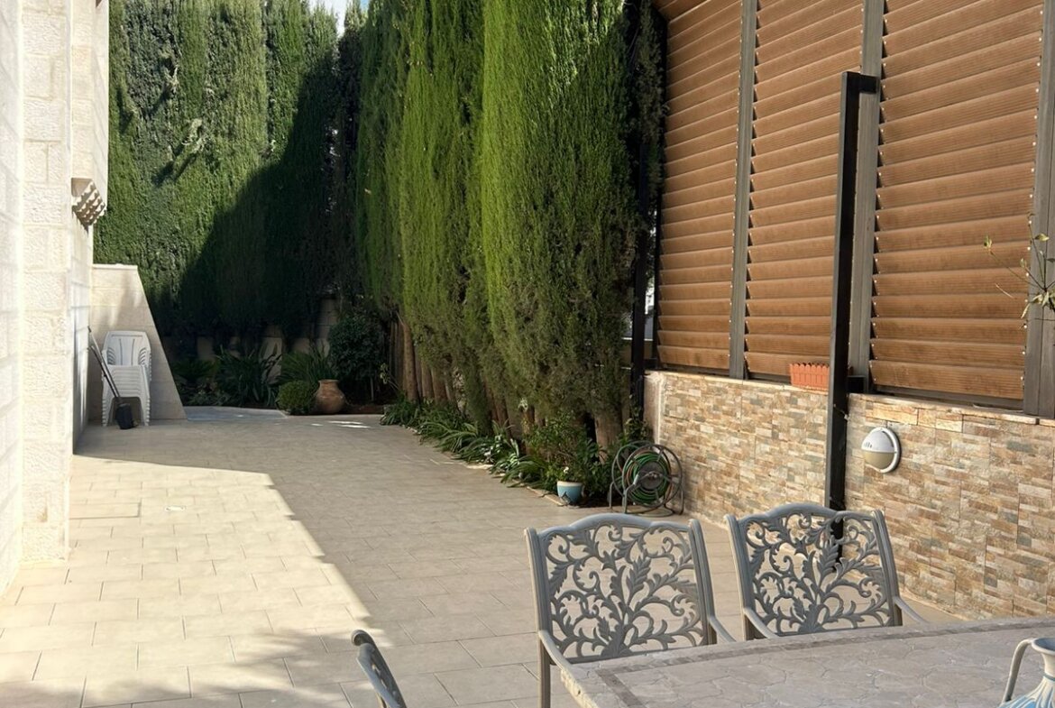 Outdoor patio with a long privacy hedge of tall evergreen trees along the left and a wood slat fence on the right, metal dining table and chairs in the foreground under a blue sky.