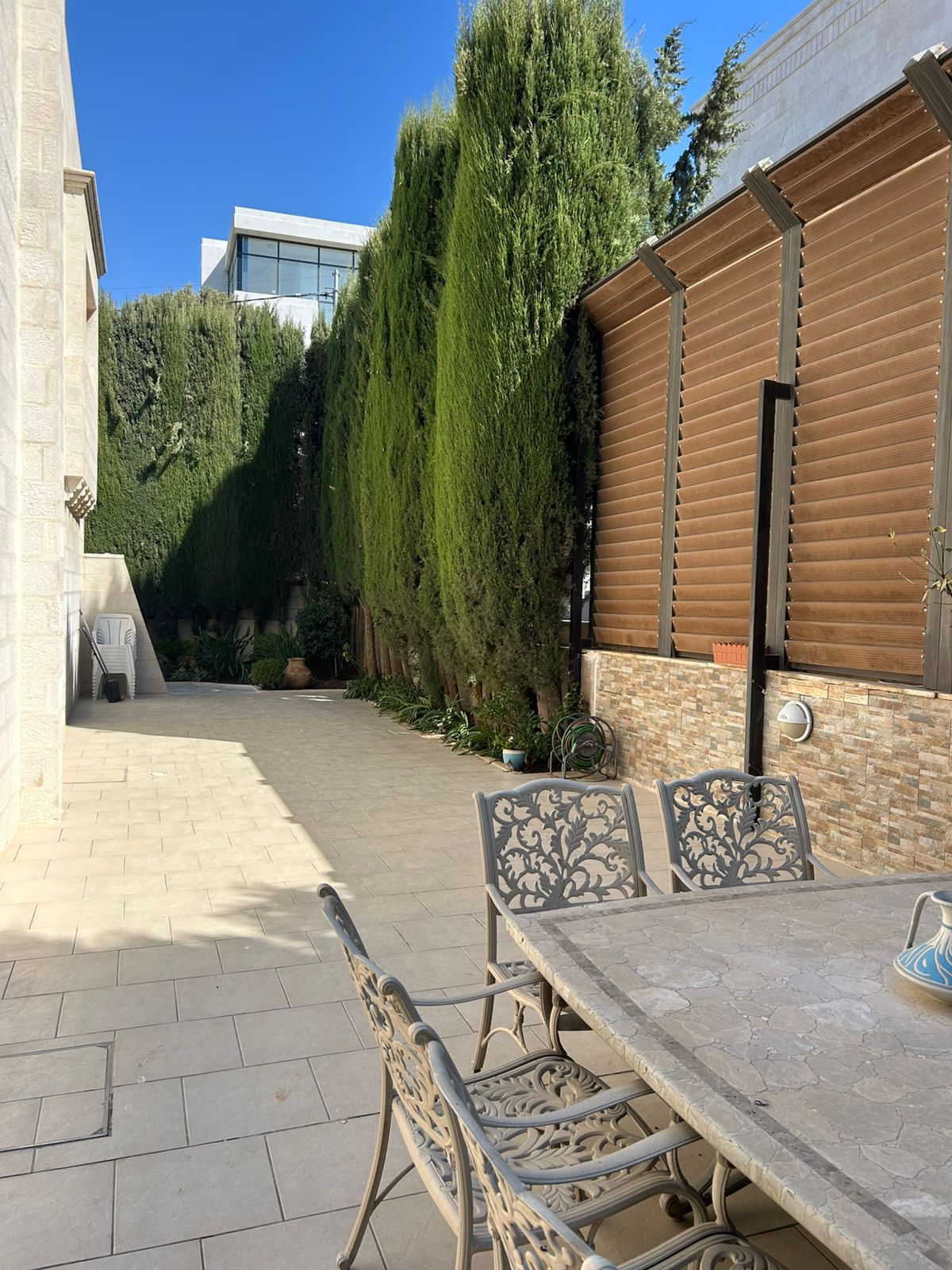 0e591716-7c32-4c3f-a191-5a03d1c76743 Outdoor patio with a long privacy hedge of tall evergreen trees along the left and a wood slat fence on the right, metal dining table and chairs in the foreground under a blue sky.