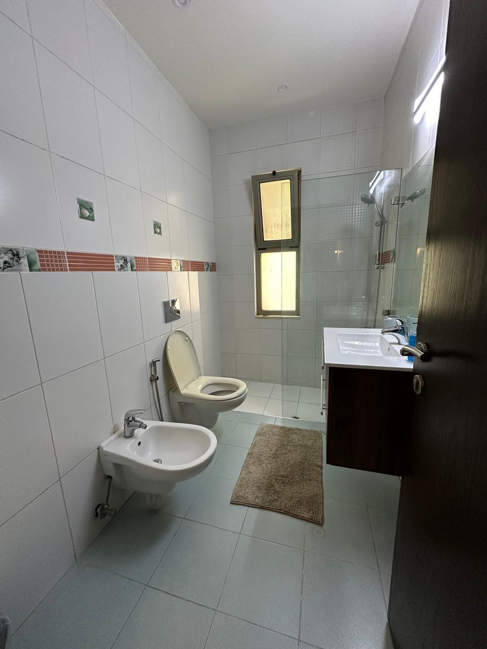 0e6ad83a-b914-4c0c-b6c3-8ca5278fa921 A modern bathroom with a wall-mounted toilet, bidet, and a white sink with dark wood cabinet beneath a glass shower enclosure.