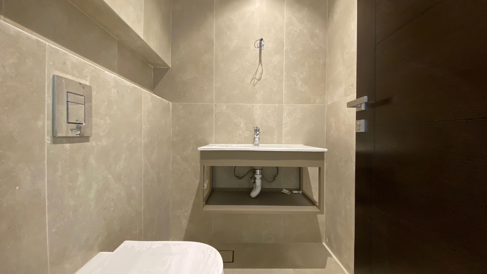 0e7fa389-325f-4f59-b00a-a62e443d8d52 Beige tiled bathroom with a wall-mounted flush plate, white sink on an open cabinet showing exposed pipes and a toilet at the bottom left; construction wires above the sink.