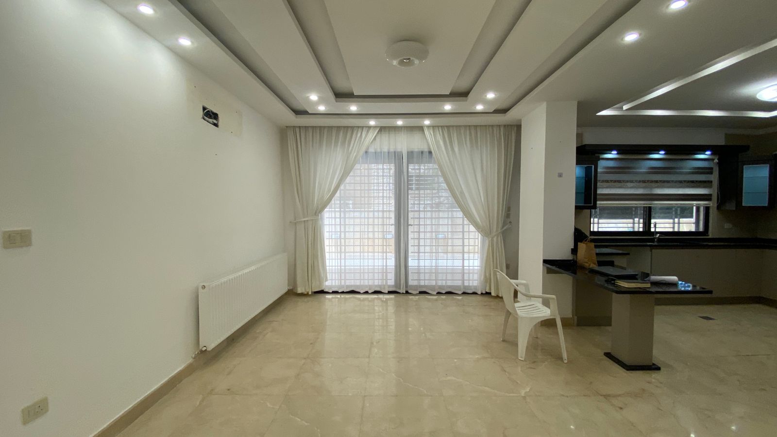 0e9f7d72-b0c3-4dc2-a702-7186db76effc Empty living area with large glass doors and sheer white curtains, modern recessed ceiling lights, and marble floor; a white chair sits by a kitchen island to the right.