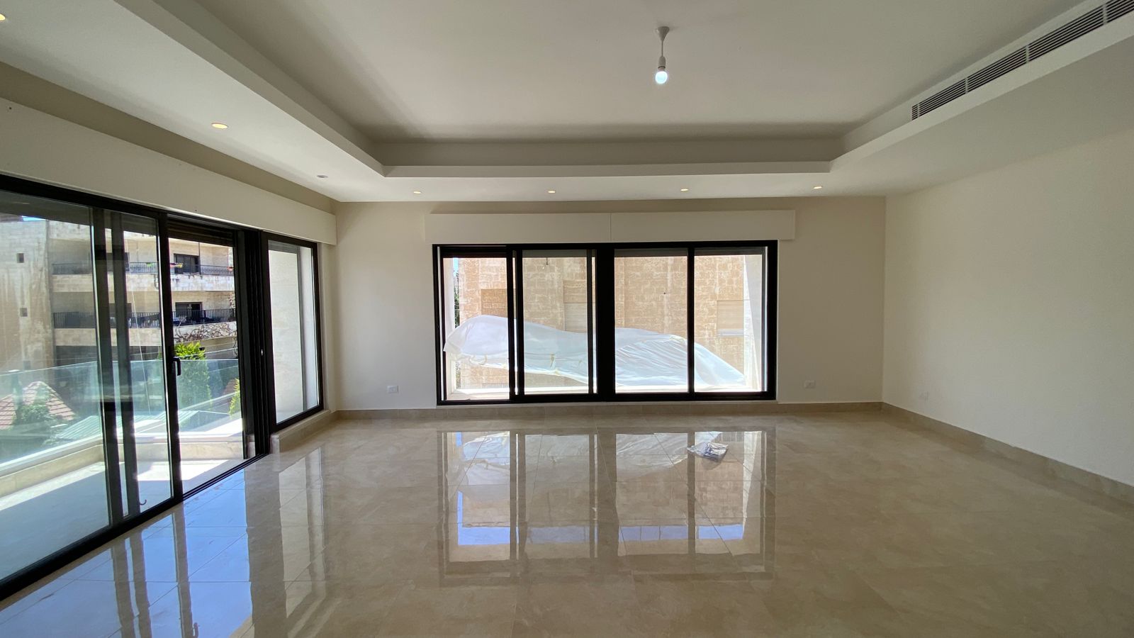 0f047d59-76fd-476a-8a03-0182239095c3 Bright empty living room with glossy beige tile floor and large sliding glass doors opening to a balcony.Parsed reflections on the floor and a minimalist ceiling with recessed lighting reveal a modern, open space.