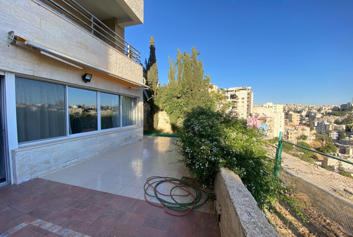 Terrace of a beige stone apartment building with sliding glass doors and a coiled garden hose on tiled flooring; city skyline in the distance under a blue sky.