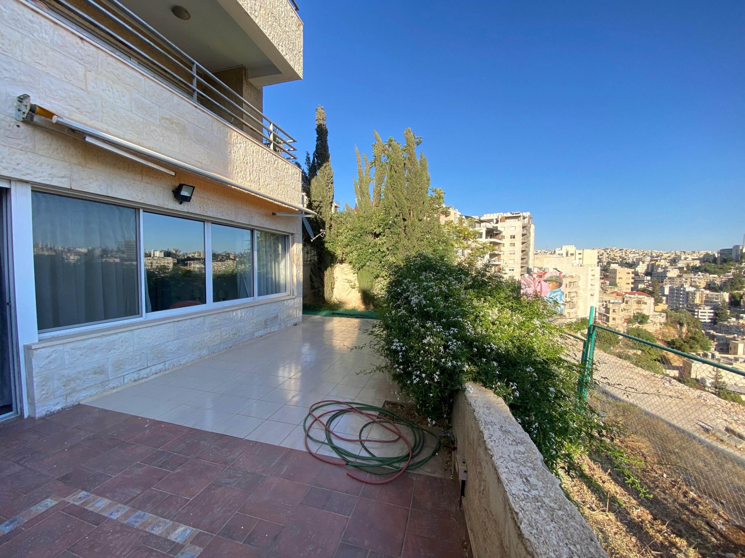 0f473536-ef42-4bc0-b8db-ab8d73cbfb9e Terrace of a beige stone apartment building with sliding glass doors and a coiled garden hose on tiled flooring; city skyline in the distance under a blue sky.