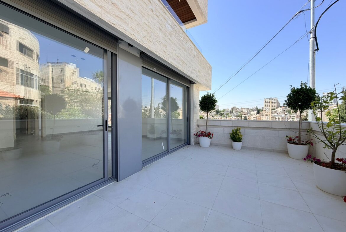 Modern balcony with large sliding glass doors, white tile floor, and potted plants overlooking a cityscape.