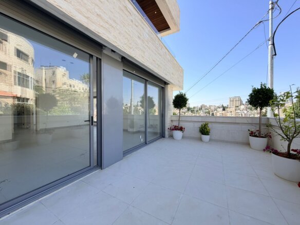 Modern balcony with large sliding glass doors, white tile floor, and potted plants overlooking a cityscape.
