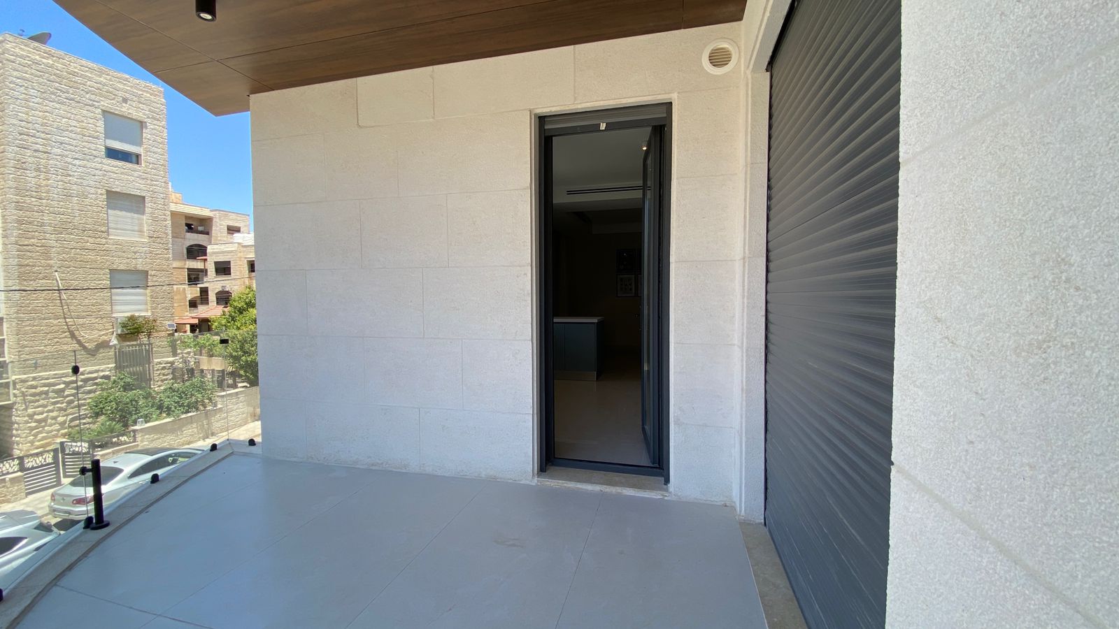 1101b5df-a97f-4b58-b09c-7da16f86a2e8 Modern balcony with glass railing overlooking a street of stone buildings; a sliding door is open to the interior.