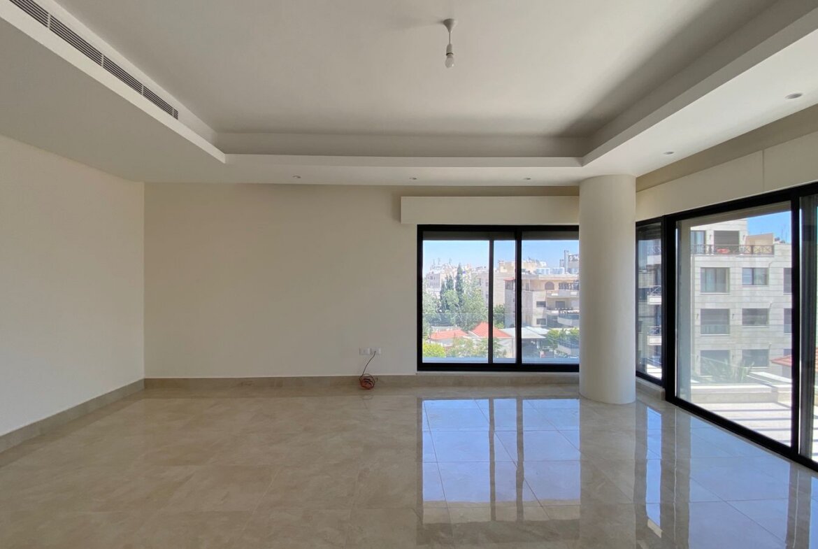 Empty modern living room with large floor-to-ceiling windows showing a city view and balcony beyond. Light-colored tiled floor and white walls with a recessed ceiling. A single exposed wire rests on the floor near the wall.