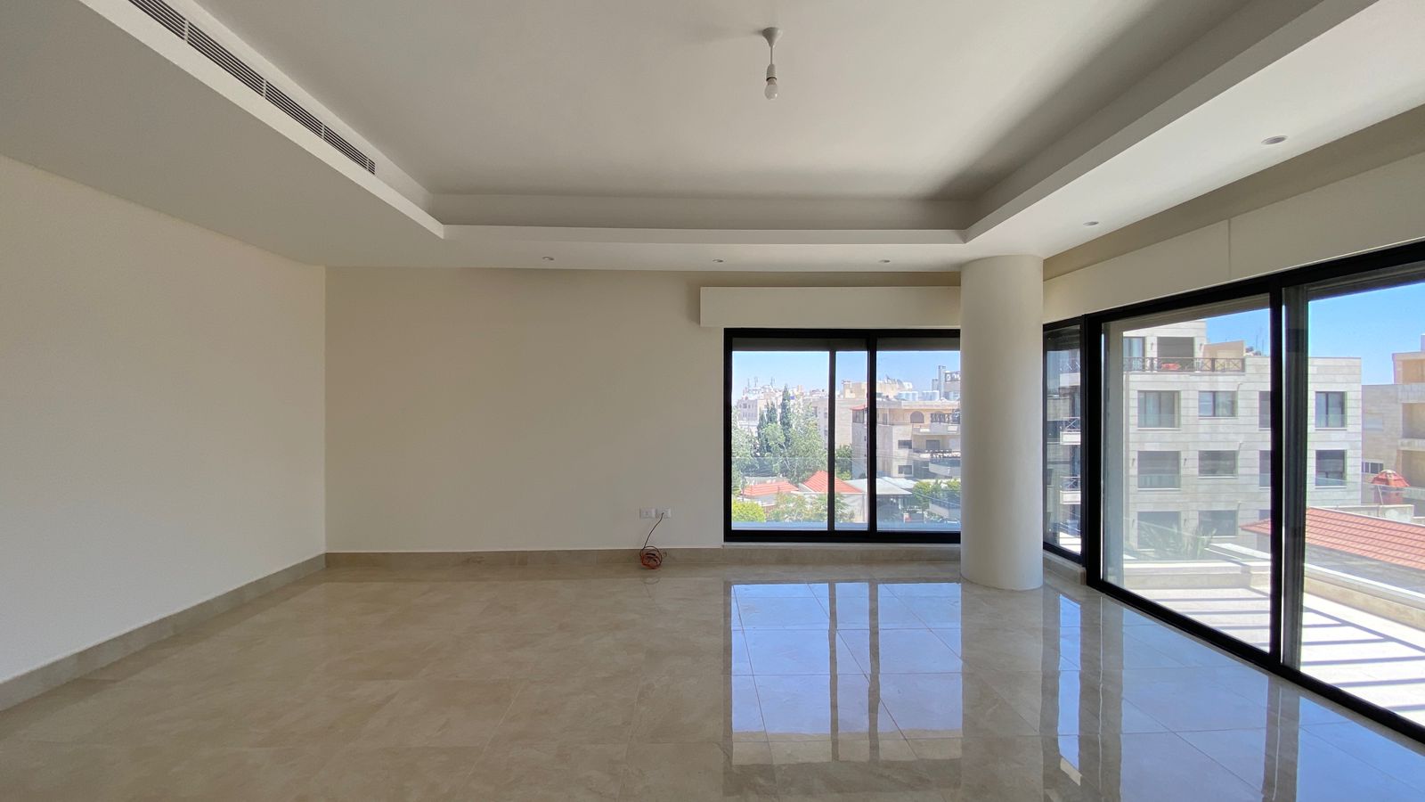 11599fe6-b437-4174-b8f1-61ab2043189a Empty modern living room with large floor-to-ceiling windows showing a city view and balcony beyond. Light-colored tiled floor and white walls with a recessed ceiling. A single exposed wire rests on the floor near the wall.