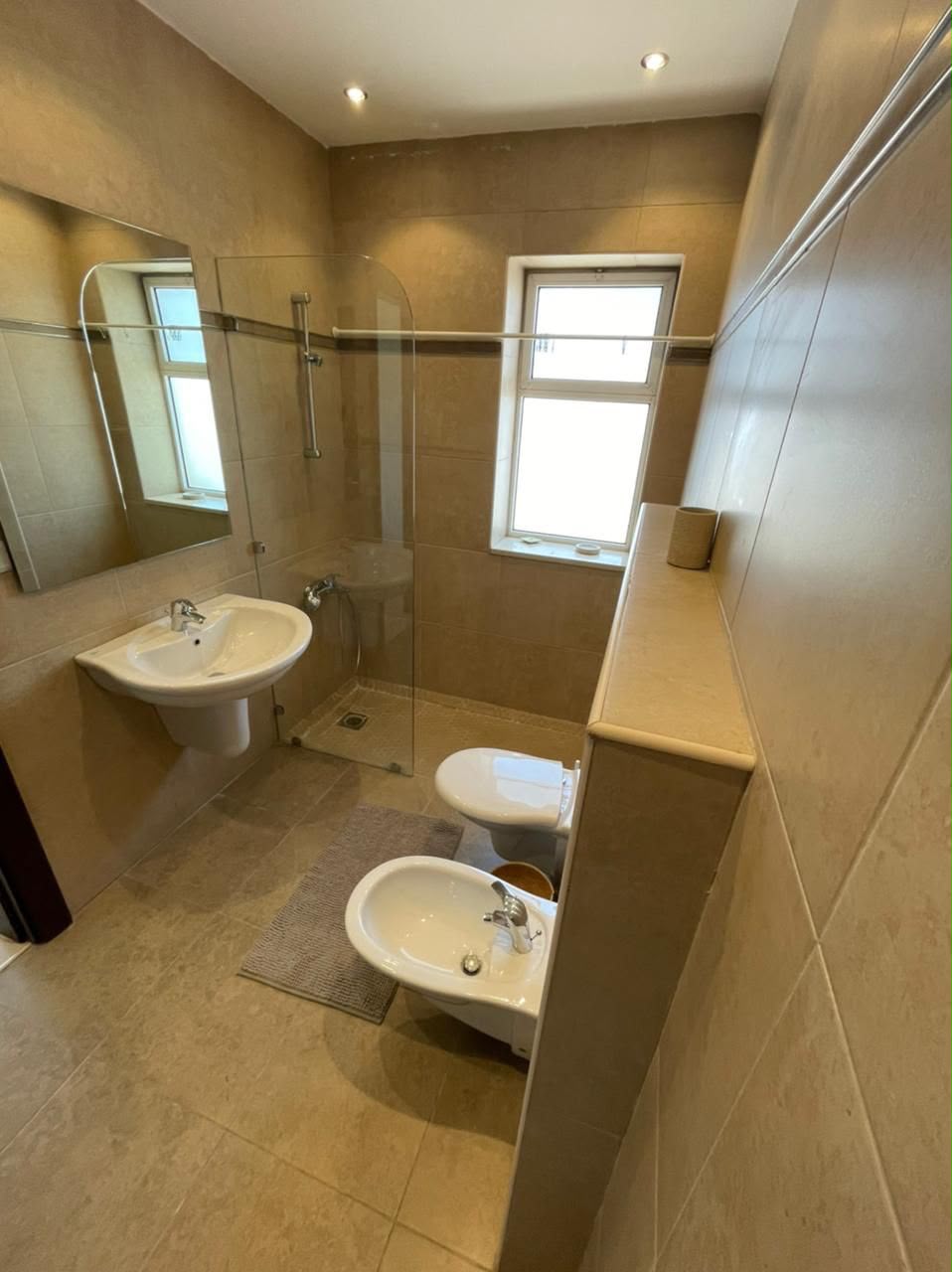 118f0b0c-b64f-4d82-ae6c-54addf39641c Contemporary beige-tiled bathroom with glass shower enclosure, wall-mounted sink, toilet and bidet, and a small window letting in light.