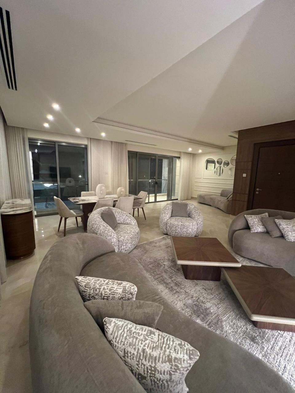 130fda95-9e2b-4df4-80af-ab1e96e6abdf Luxurious open-concept living room with a curved gray sectional sofa, patterned cushions, two matching round chairs, and a wooden coffee table, overlooking a balcony through glass doors with sheer curtains.
