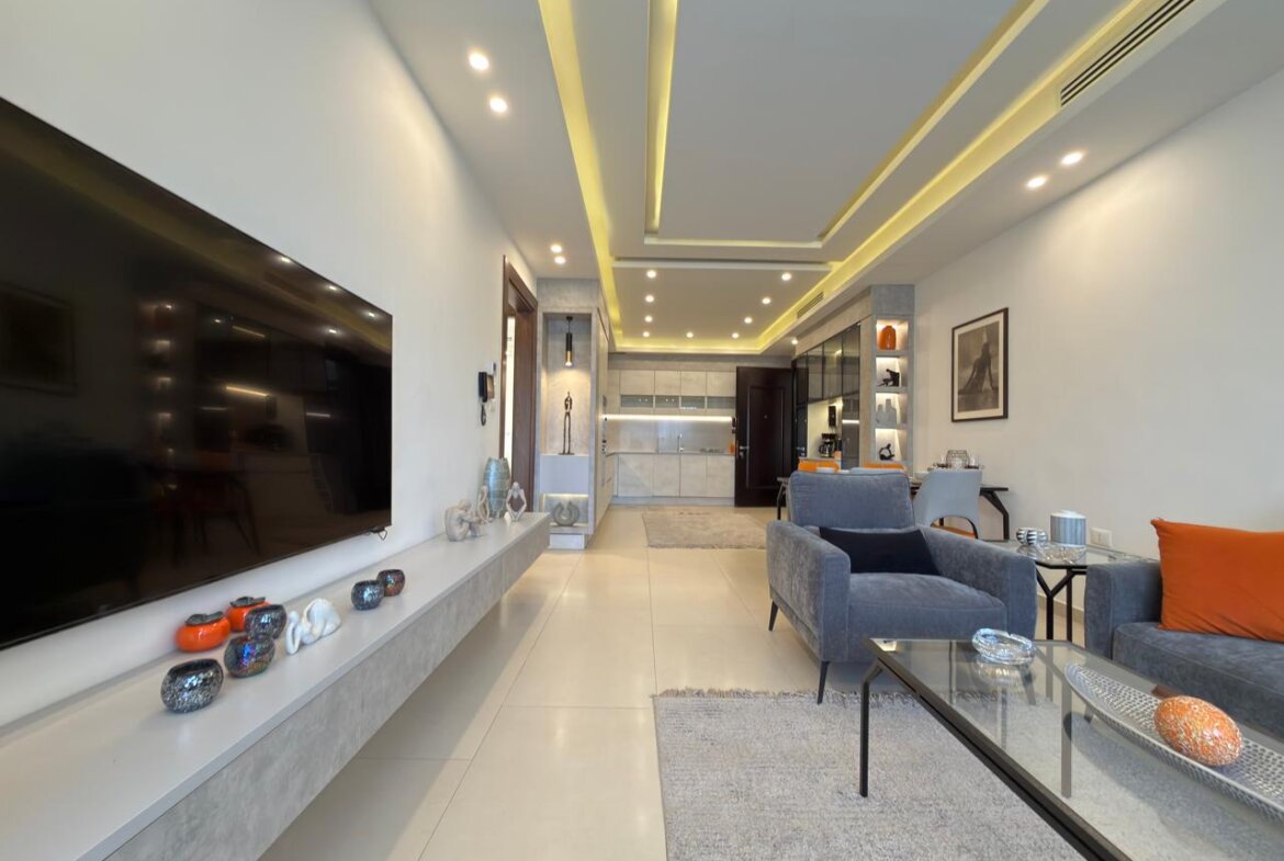 Open-plan living room with a wall-mounted TV, gray seating, orange pillow, and a glass coffee table.