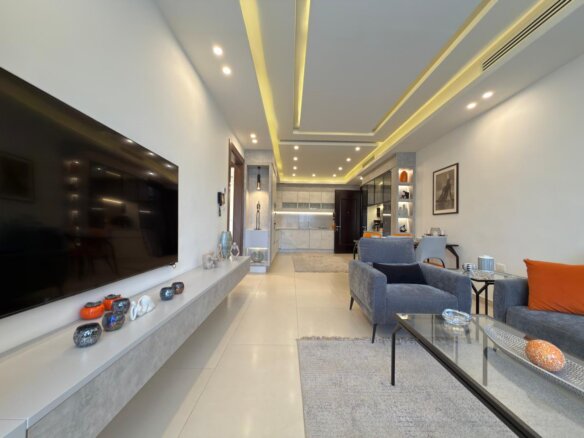 Open-plan living room with a wall-mounted TV, gray seating, orange pillow, and a glass coffee table.
