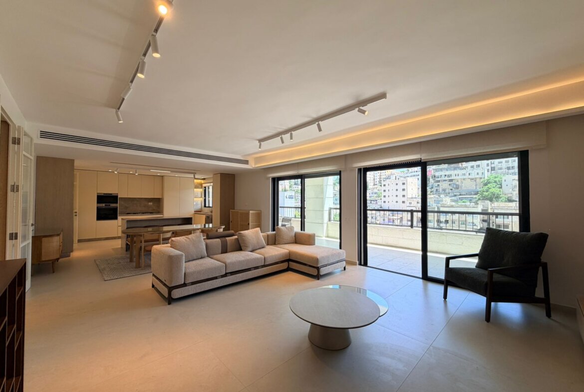 Open-plan living area with a beige L-shaped sofa, round coffee table, and large sliding doors to a balcony.