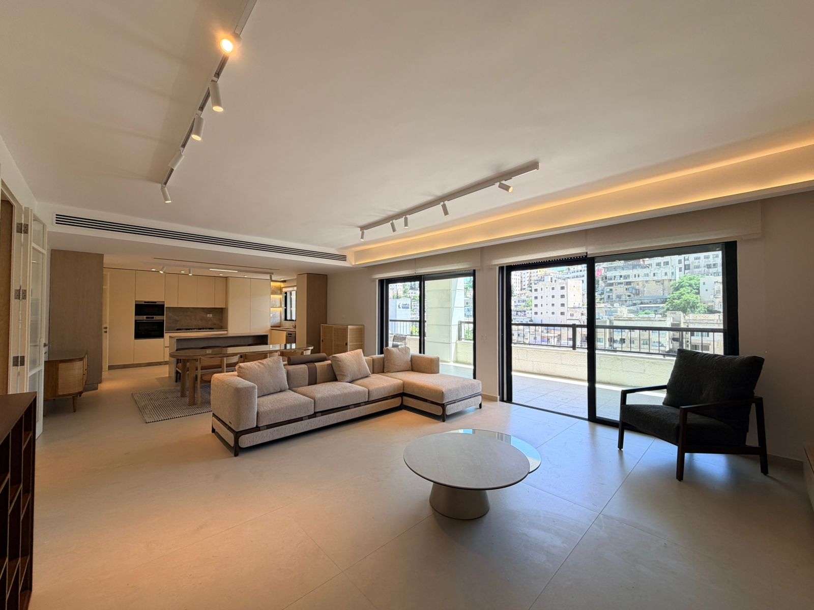 13defb1d-e1c5-41a8-9286-3ea557553ba3 Open-plan living area with a beige L-shaped sofa, round coffee table, and large sliding doors to a balcony.