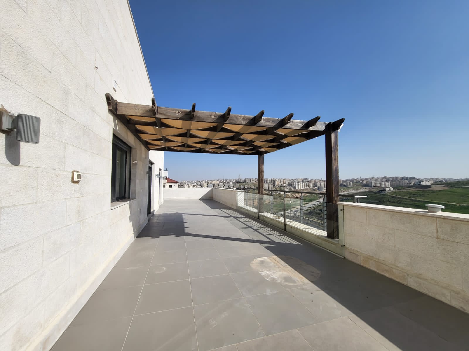 1471db0a-4b65-496b-9290-ac63b0d3cc19 Rooftop terrace with a brown wooden pergola, glass railing, and a distant city skyline under a clear blue sky.
