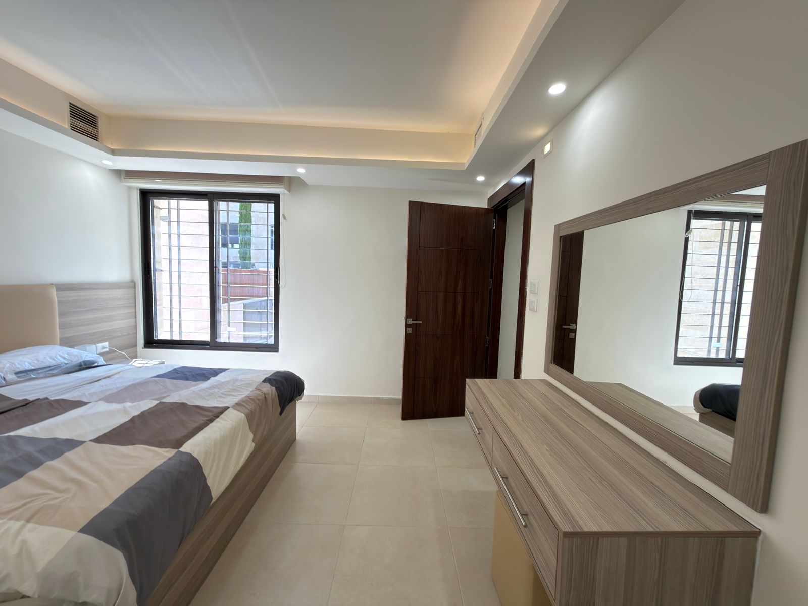 14c95e71-728d-4843-a2c1-638b70f15ae7 Modern bedroom with a low wooden bed, striped gray-and-white duvet, and a large dresser with a wall-mounted mirror nearby.