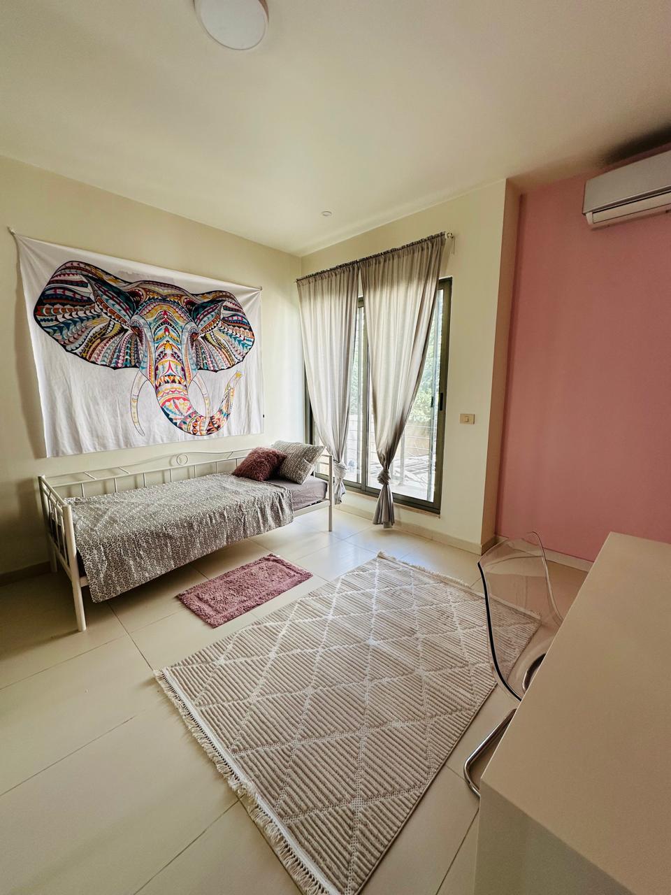 15264ab5-992b-4a23-a101-60f6645a4f01 Cozy bedroom with a white metal single bed, colorful elephant tapestry on the wall, pale curtains, and a geometric rug.