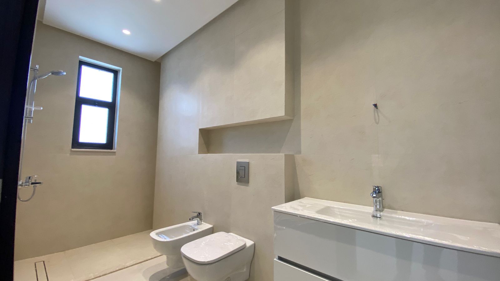 154642d3-5e6e-46f5-a940-7c3750b3be23 Modern beige bathroom with toilet, bidet, white vanity, recessed shower, and tall window.