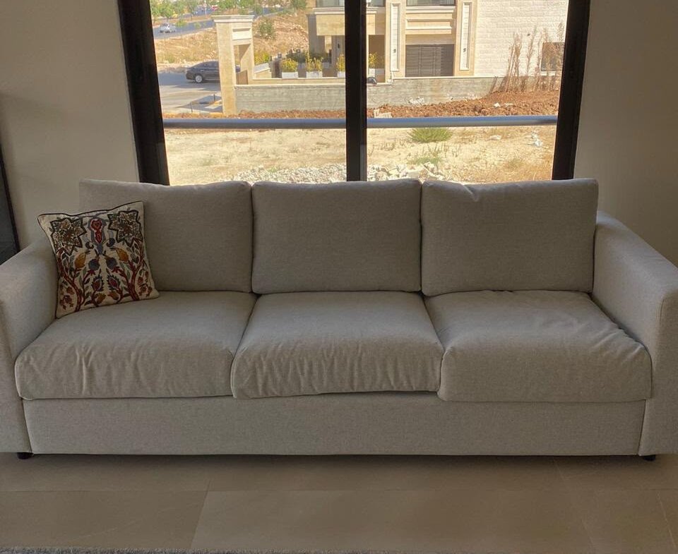 Cream-colored three-seat sofa with a patterned decorative pillow, set before a large window overlooking buildings and a hillside outside.