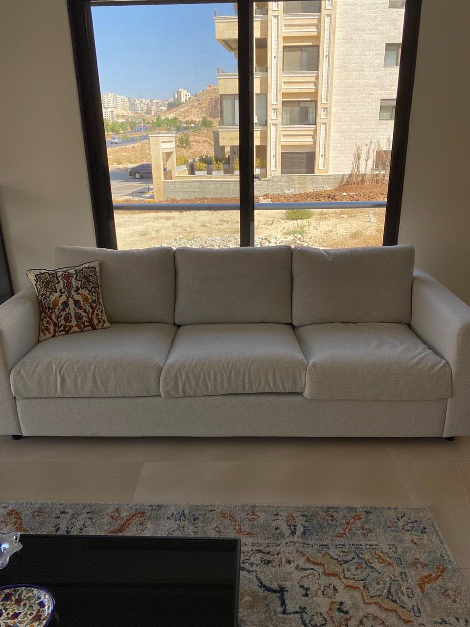 18900a60-349e-4a92-a37f-ce8583f0a9ae Cream-colored three-seat sofa with a patterned decorative pillow, set before a large window overlooking buildings and a hillside outside.