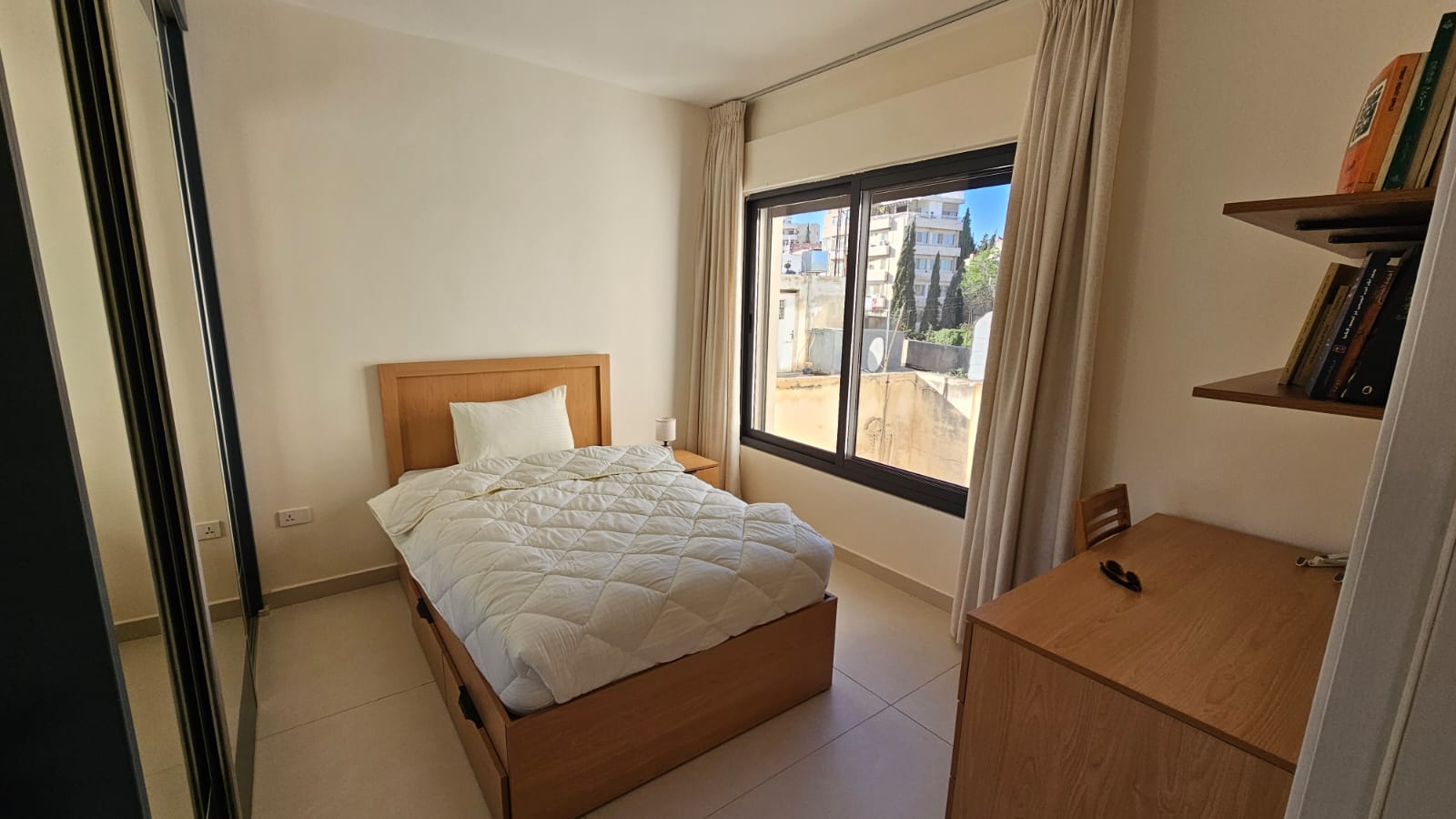 19826970-d1f0-4940-8bf5-5dbd7ab6830c Small bedroom with a wooden bed, white bedding, and a nightstand beside a large window with beige curtains