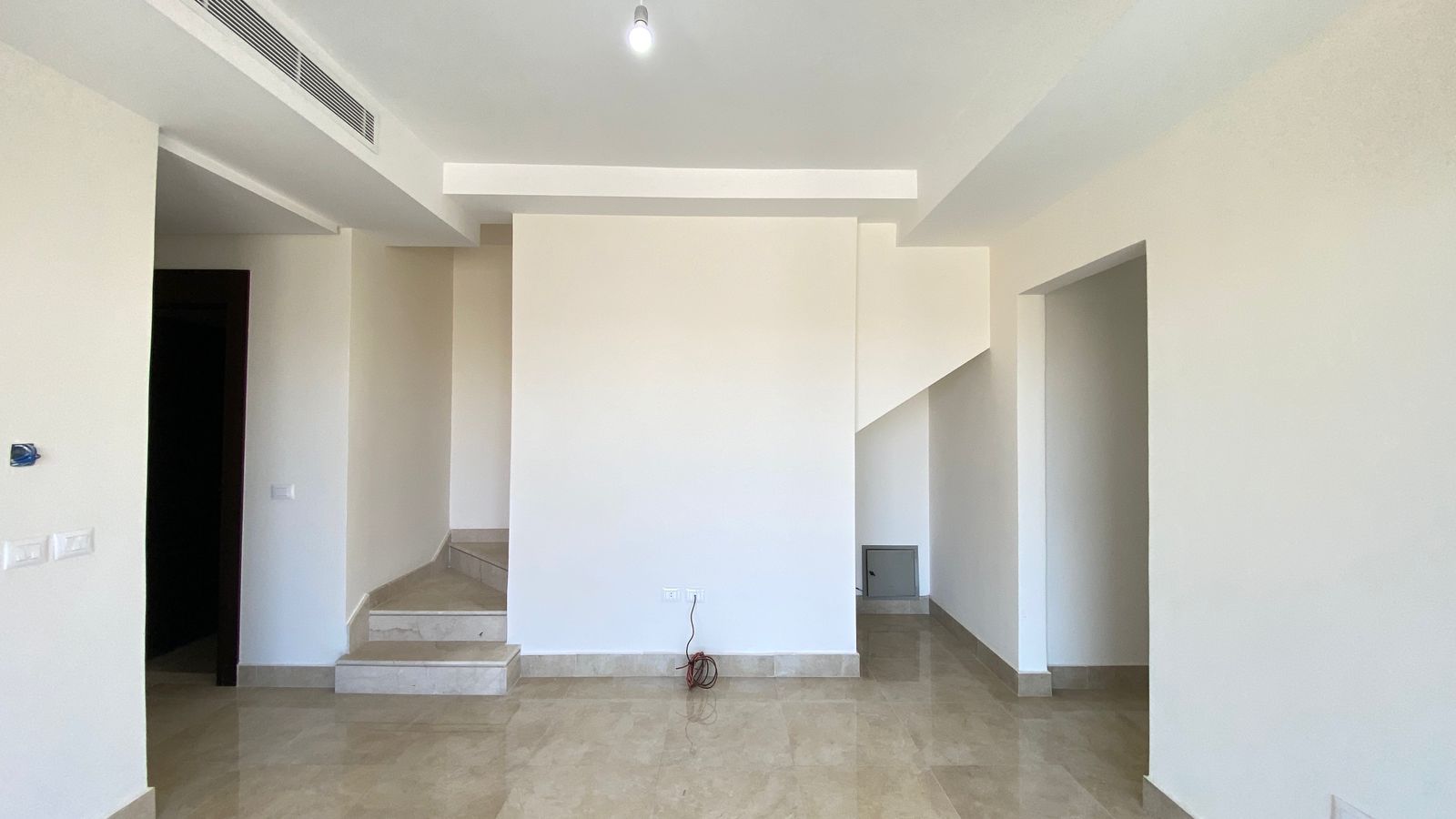 1992a4a2-4b81-4b1a-be80-9dceeb98a510 Empty modern living space with beige tile floor, white walls, a central column, and staircase to the left.