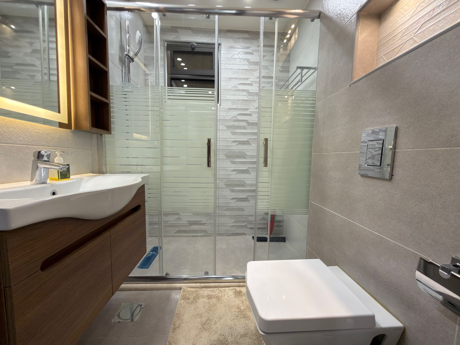 19a077a3-06b5-4295-b582-9c5bfa09f55a Contemporary bathroom featuring a glass shower enclosure with frosted stripes, a wall-mounted sink on a wooden vanity, and a wall-mounted toilet nearby.