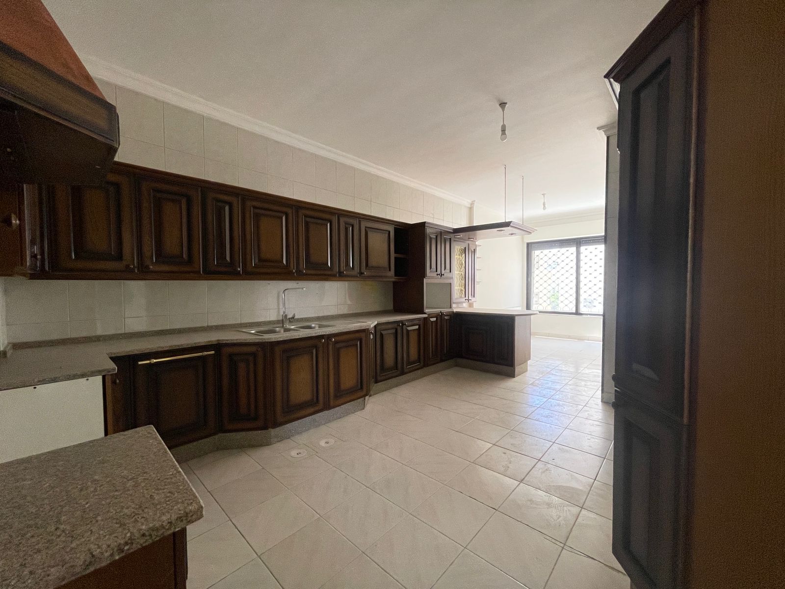 1a089bad-f583-4118-b812-3314276193a5 Kitchen with dark wooden cabinets, light tiled walls, and a central island under a bare light fixture laid out along a bright windowed wall.