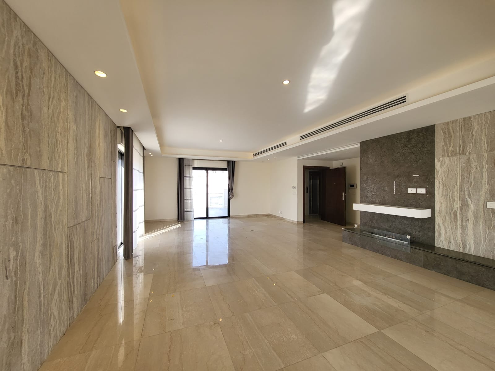 1a7ecce9-a18c-4d7b-81a4-1d6b52375dfa Spacious modern living room with marble floors, a dark stone accent wall with a floating shelf, and balcony doors with curtains.