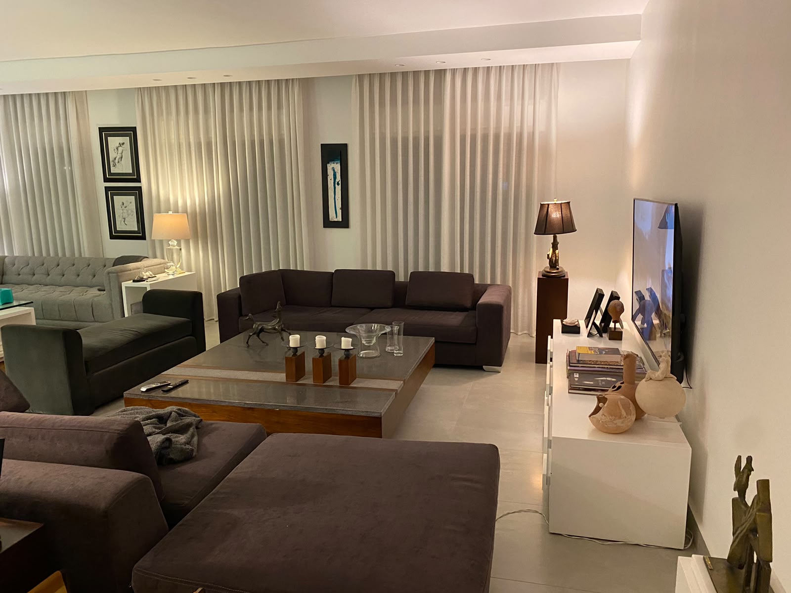 1b603250-dd88-4ab0-8b8d-96446ee53620 Modern living room with brown sectional sofas, glass coffee table, and a large flat-screen TV on a white console. Curtains span the back wall with warm lamps for ambience.