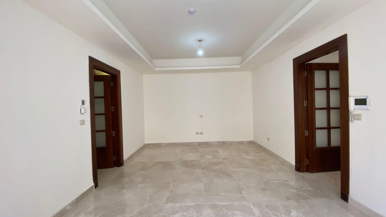 1b8405e5-c6c1-41c0-97ff-fdcbb72b5935 Empty living room with beige marble tile floor, white walls, and dark wood-framed glass doors on both sides; single ceiling light in center and a simple recessed ceiling edge.