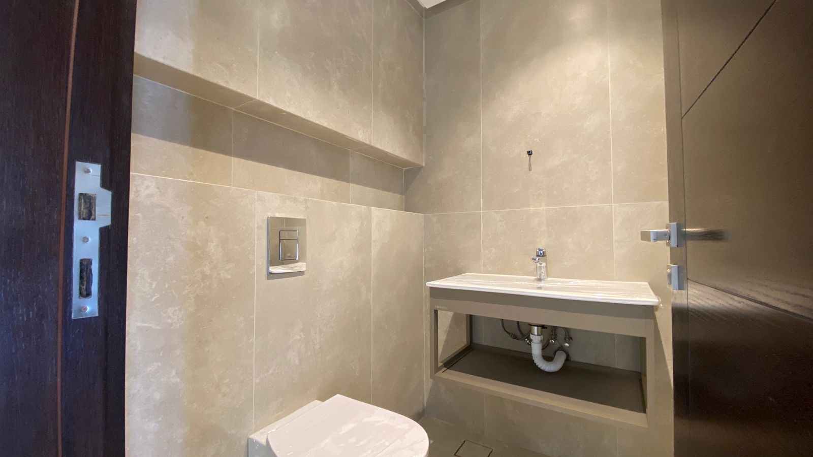1b8a1a10-7fb4-431f-9a69-752c23b1003d Modern beige-tiled bathroom with a wall-mounted toilet flush plate and a sink set in an open cabinet with exposed plumbing under a recessed shelf on a dark doorway side.