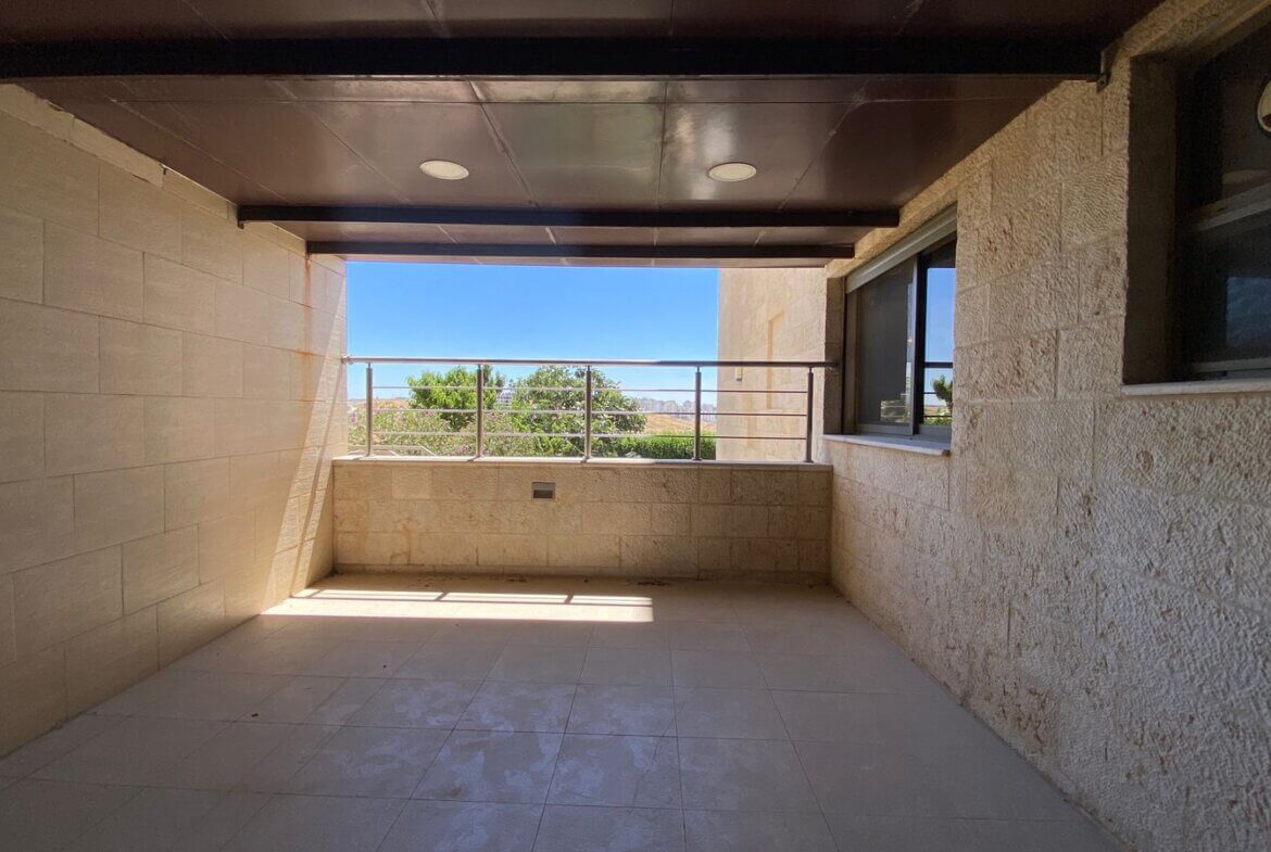 1bedf4f3-1629-4a97-9c04-bc61f655136d Covered balcony with beige stone walls and tiled floor, metal railing, overlooking trees and a blue sky beyond.