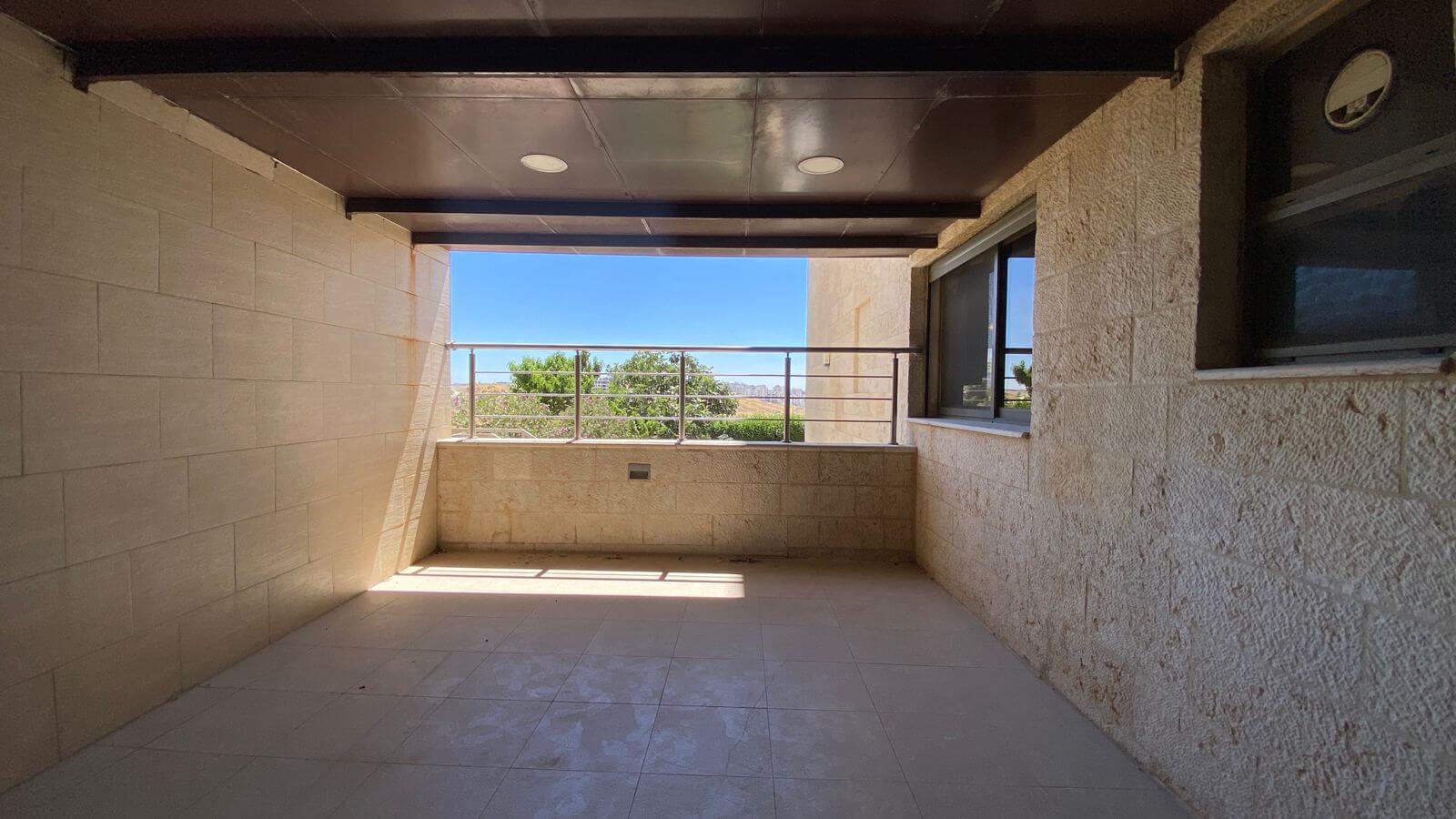 1bedf4f3-1629-4a97-9c04-bc61f655136d Covered balcony with beige stone walls and tiled floor, metal railing, overlooking trees and a blue sky beyond.