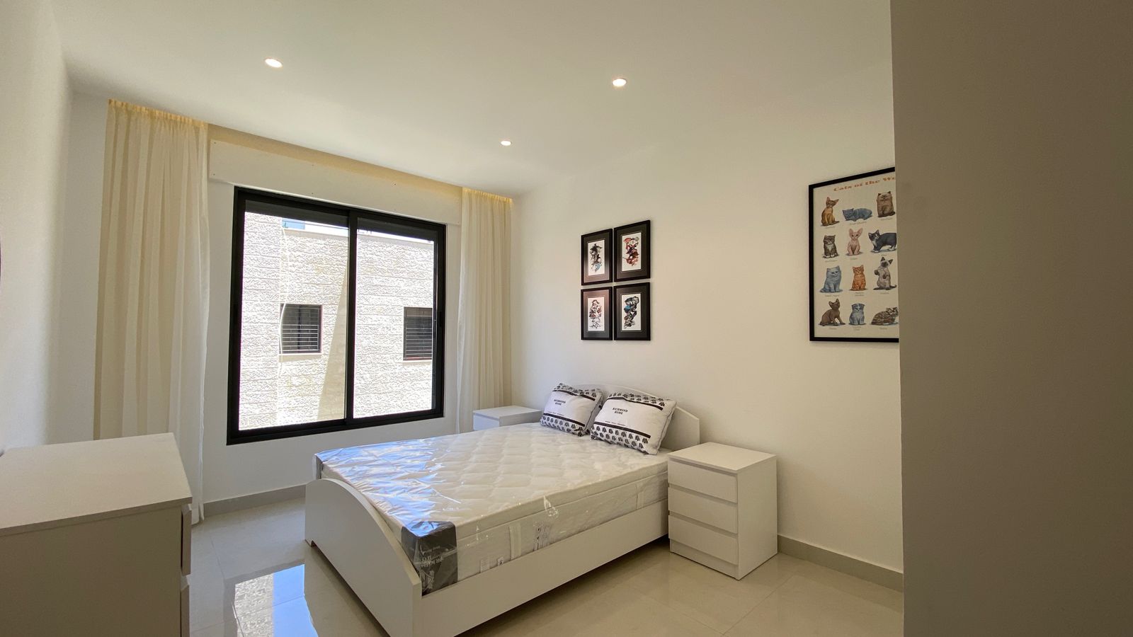 1c48ef2b-dece-4cfd-92f9-910cefc25fda Bright white bedroom with a large black-framed window, white bed, and matching nightstands; wall art of cats above the bed.
