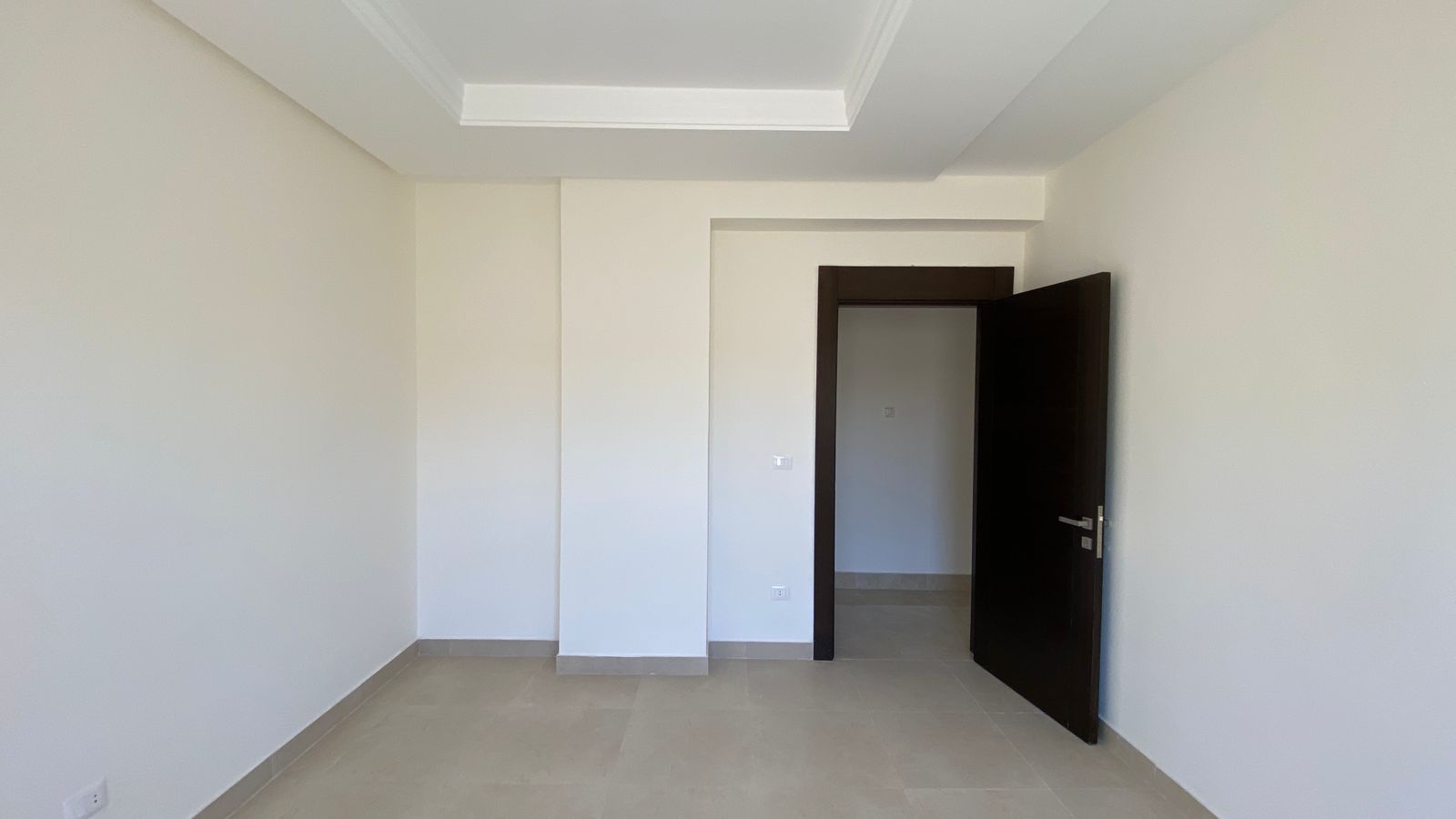 1de87ae3-e57f-4164-bc5d-51d14fb35b66 Empty room with beige floor tiles, white walls, and a dark wood door partially open to a hallway.