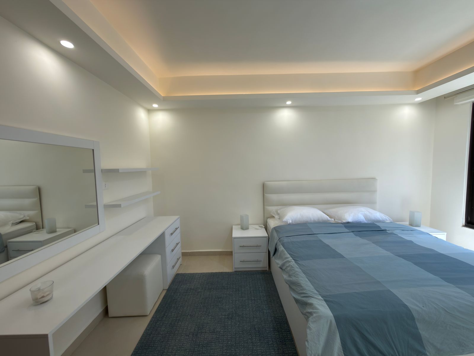 1e49db75-5040-49a5-b6fc-6d3f01b7835d Minimalist white bedroom with a blue-gray striped comforter, white headboard, and matching nightstands.