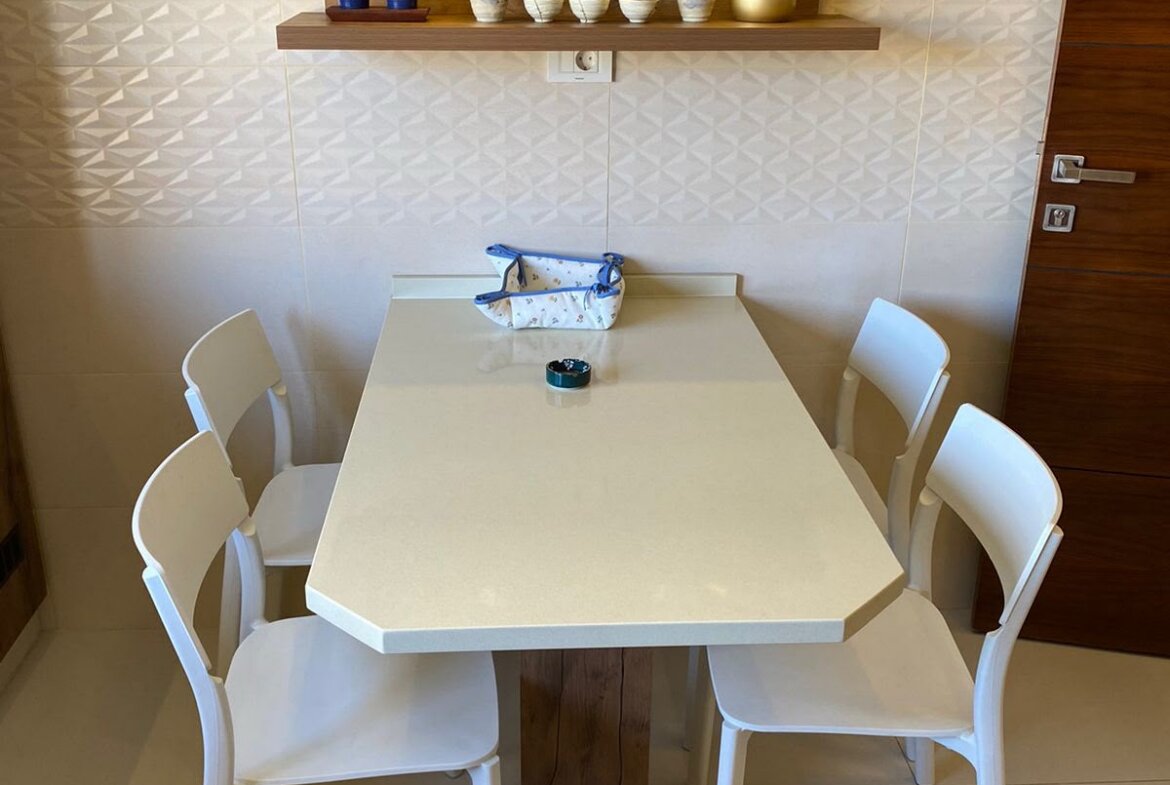 White hexagonal dining table with six white chairs in a bright kitchen nook; a wooden shelf above holds teacups and a yellow kettle, with a blue patterned bag on the table.