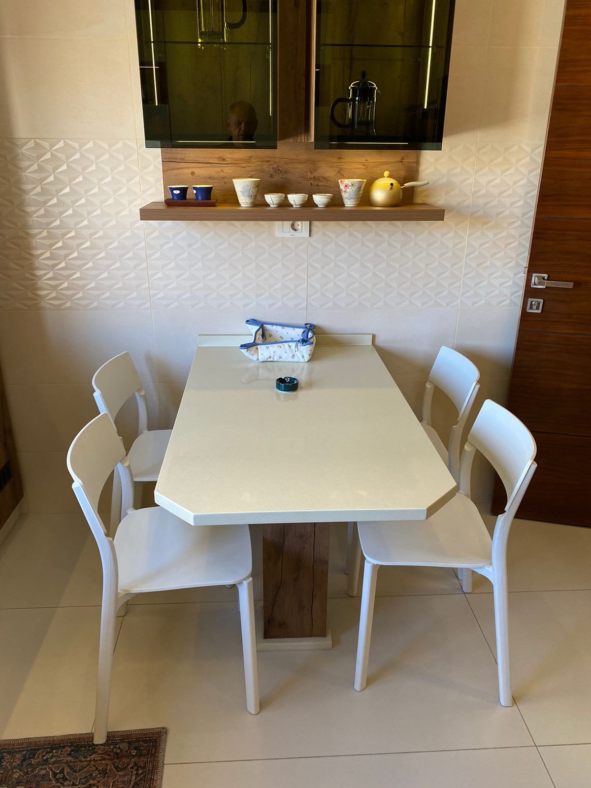 1ee4c557-fce5-4a59-9f08-5293c809db99 White hexagonal dining table with six white chairs in a bright kitchen nook; a wooden shelf above holds teacups and a yellow kettle, with a blue patterned bag on the table.