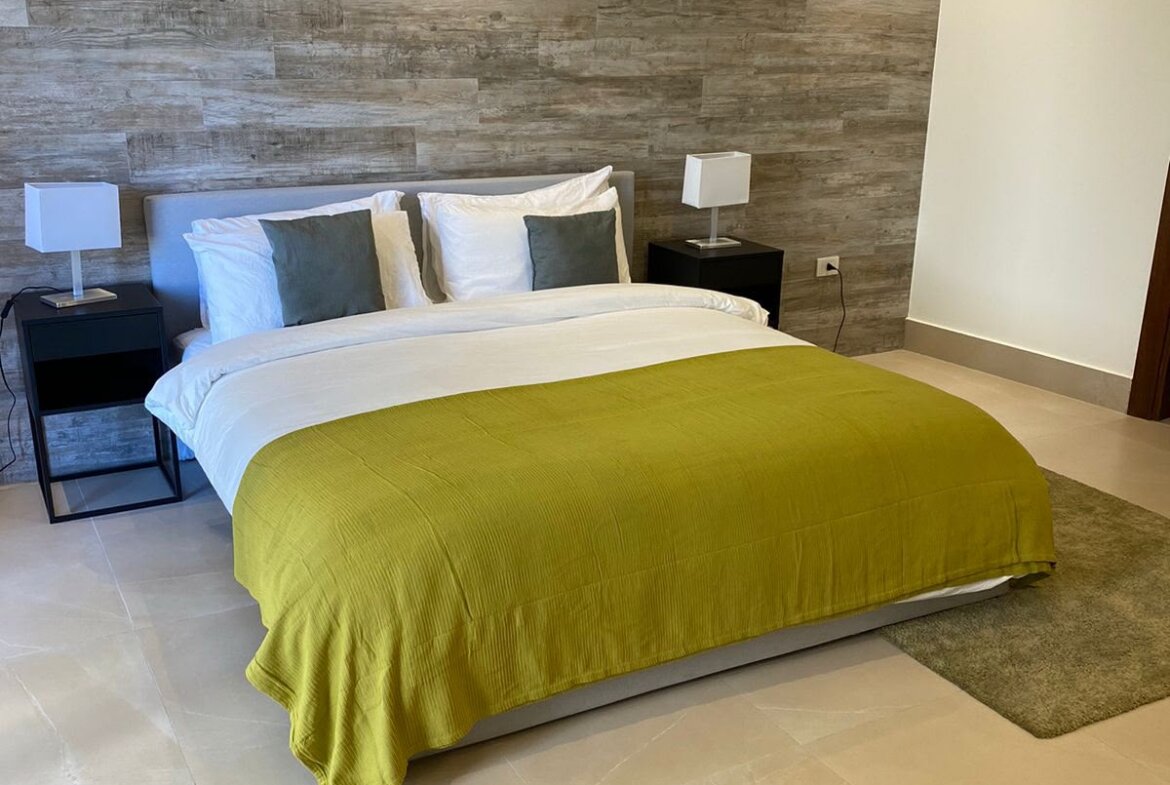 Modern hotel-style bedroom with a king bed, white linens, lime-green duvet, gray pillows, flanked by black nightstands and square lamps, wood accent wall behind.