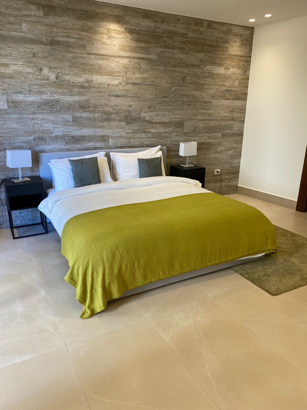 20d0fdc5-5d38-4997-9906-35d300bfec2e Modern hotel-style bedroom with a king bed, white linens, lime-green duvet, gray pillows, flanked by black nightstands and square lamps, wood accent wall behind.