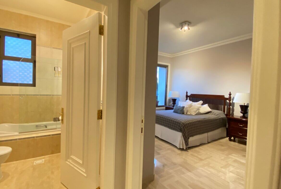 2346eb14-9bd2-4300-ad32-0fbd52172ddc View down a hallway with an open bathroom door to the left (bathtub visible) and a bedroom door to the right showing a wooden bed and two nightstands with lamps.